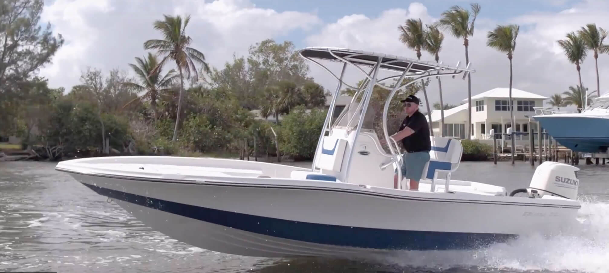 A man drives a white and blue motorboat with a T-top canopy on a calm, sunny waterway with palm trees and houses in the background.