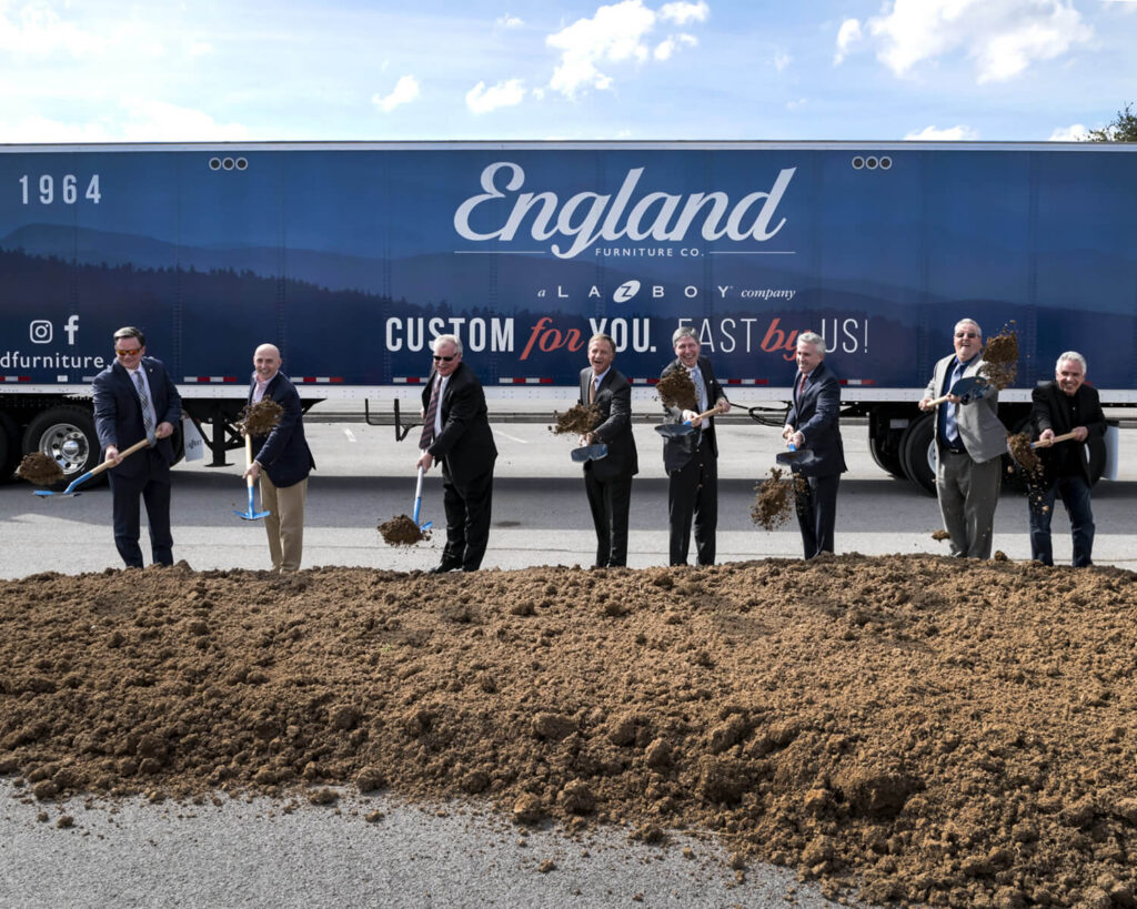 Eight men in suits stand in front of a large England Furniture Co. truck, each holding a shovel and tossing dirt in a groundbreaking ceremony. A mound of soil is in front of them under a partly cloudy sky.