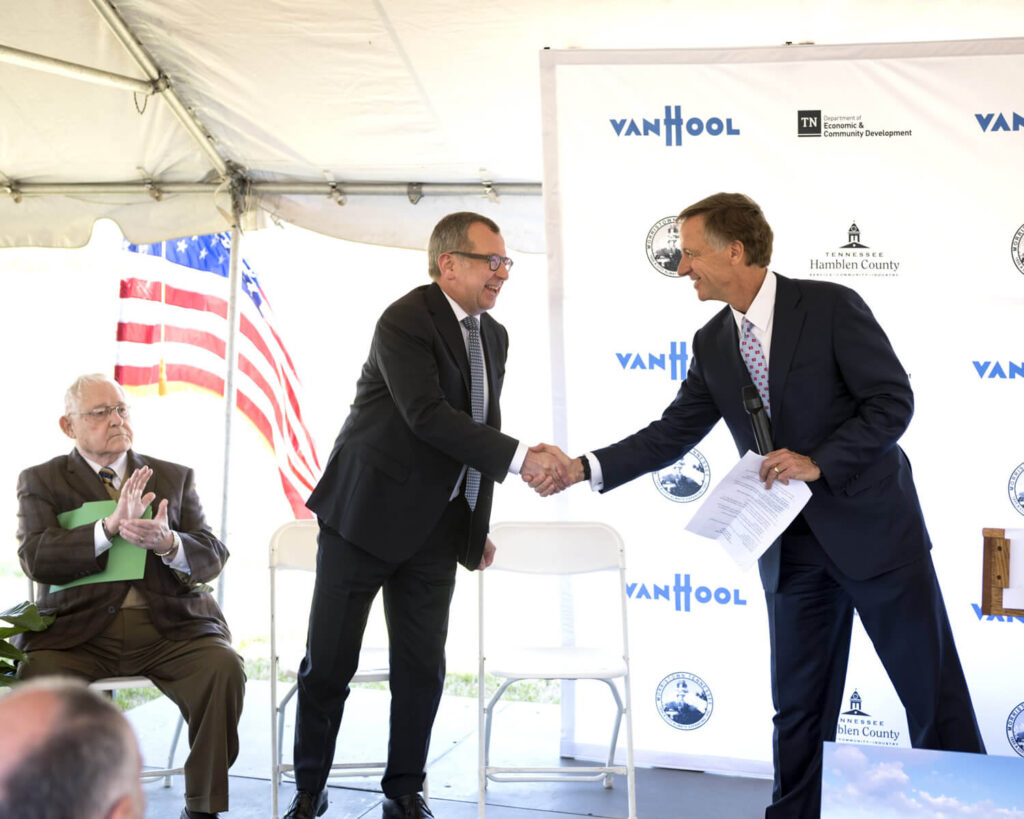 Three men at an outdoor event, two standing and shaking hands while smiling, one seated and clapping. An American flag and a backdrop with logos, including Van Hool, are visible behind them.