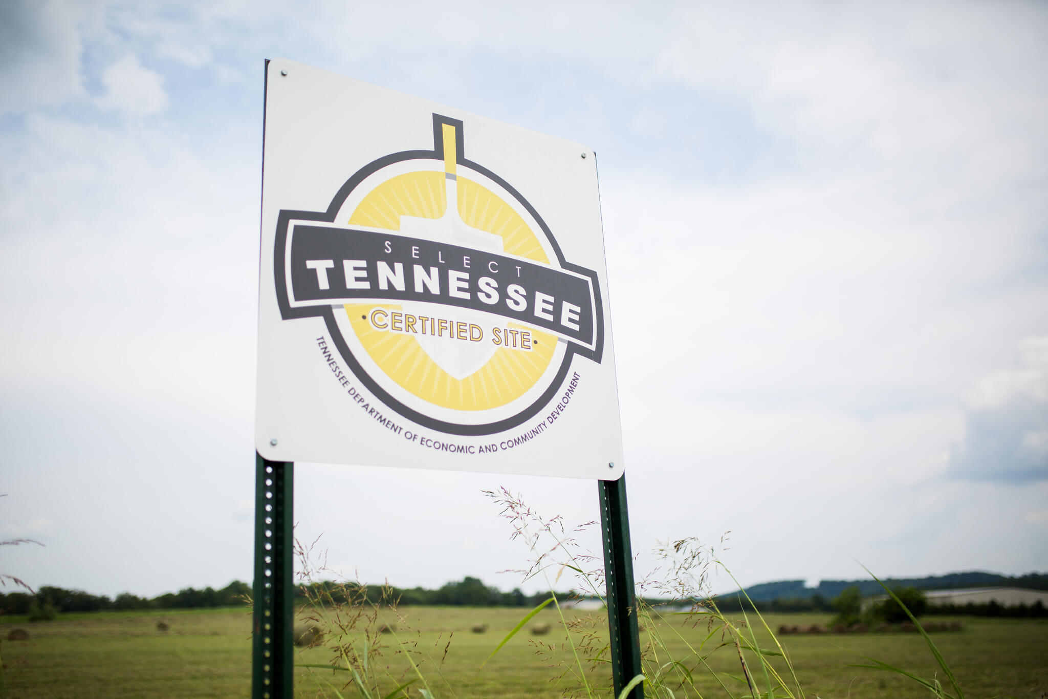 A sign in a grassy field reads "Select Tennessee Certified Site" with the Tennessee Department of Economic and Community Development seal. The sky is partly cloudy in the background.