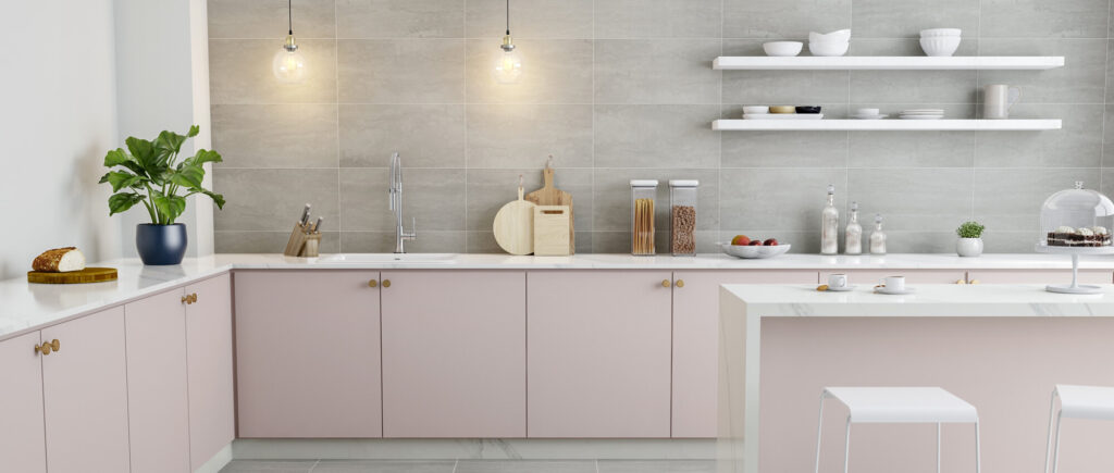 Modern kitchen with light pink cabinets, marble countertops, hanging pendant lights, open shelves holding dishes, and a kitchen island with two white stools. A plant, cutting boards, and food items are neatly arranged.