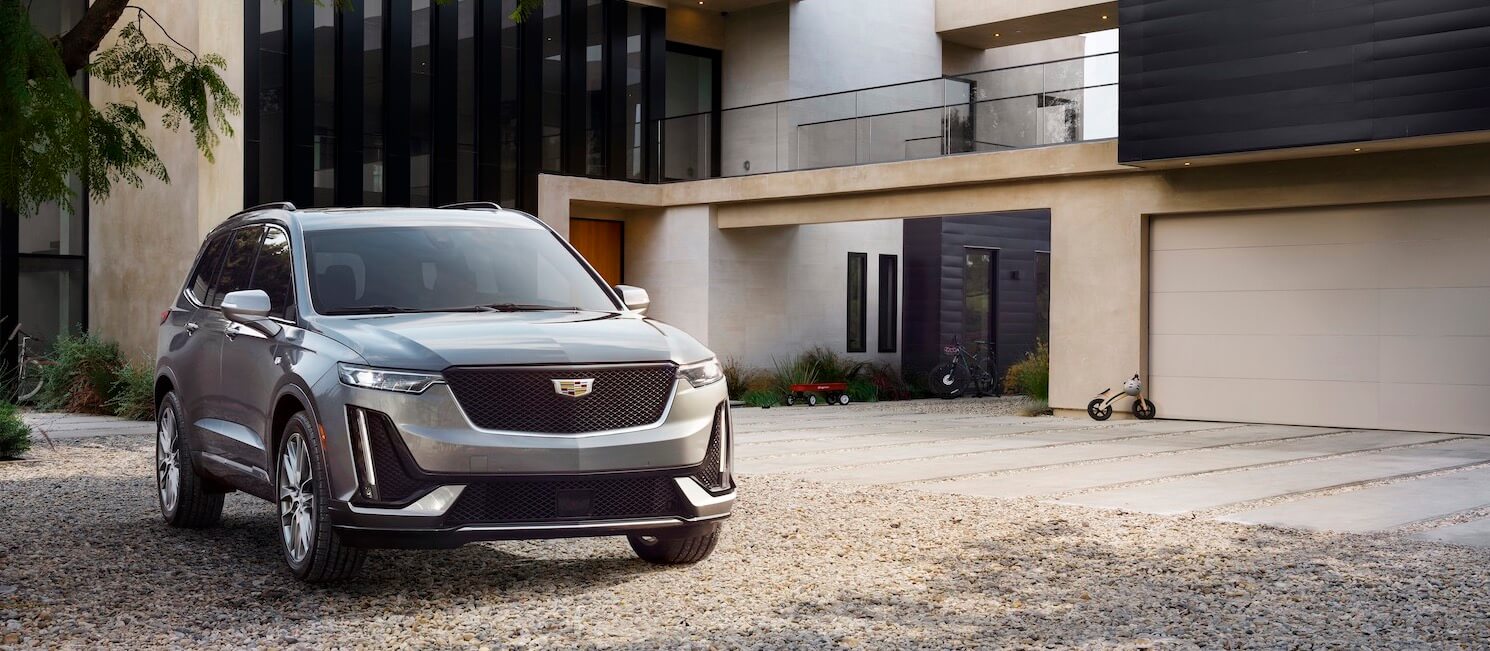 A silver Cadillac SUV is parked on a gravel driveway in front of a modern house with large windows and a garage. A small tricycle is visible near the garage door.