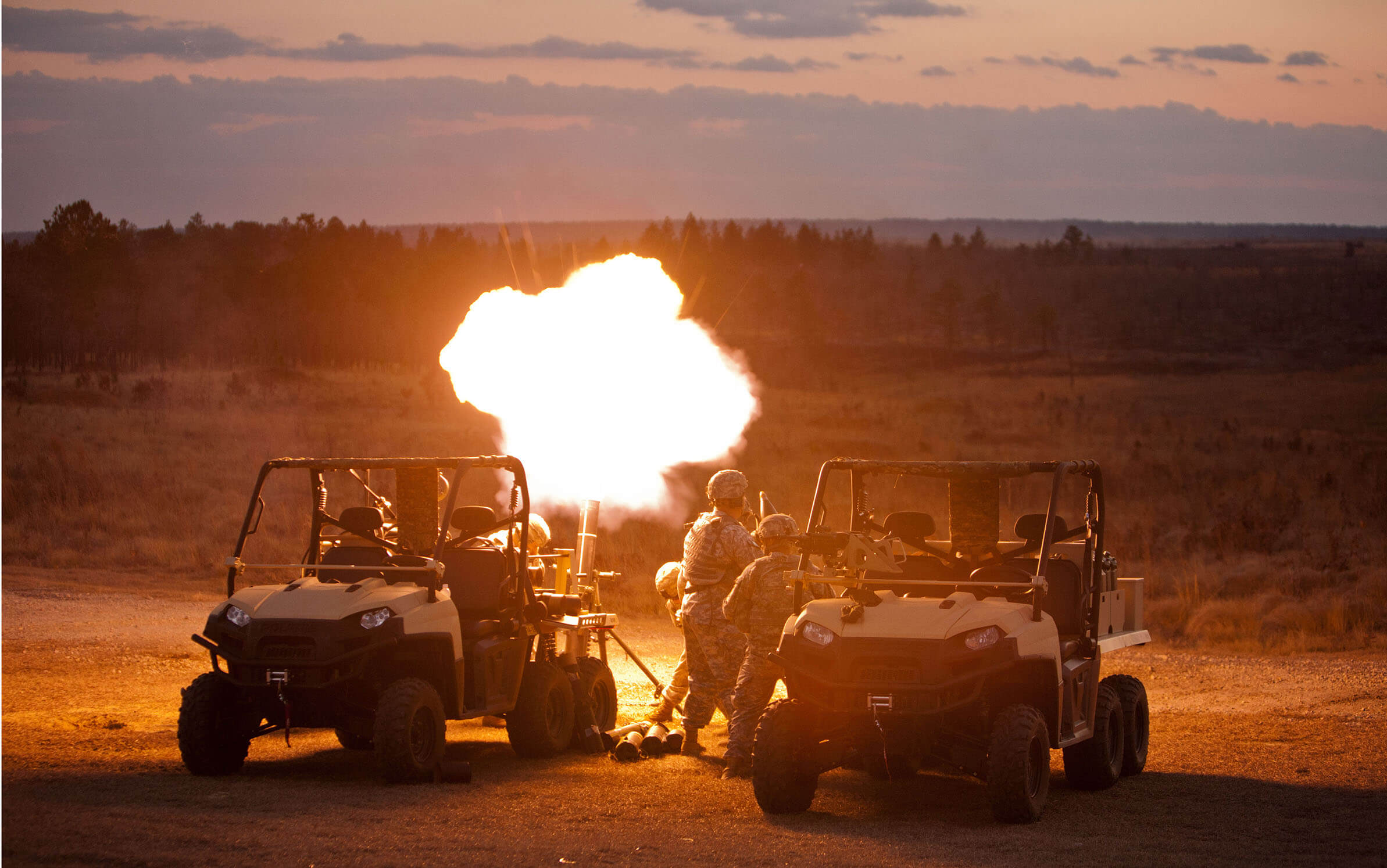 Two soldiers stand between two military utility vehicles at sunset, firing a mortar that emits a bright flash, illuminating the scene against a rural landscape.