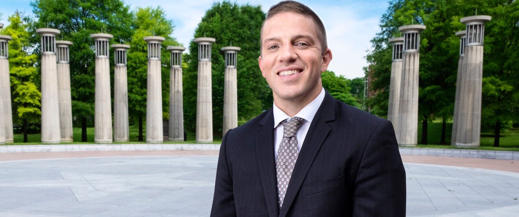 A man in a dark suit and patterned tie smiles outdoors in front of tall, evenly spaced stone pillars and green trees under a blue sky.