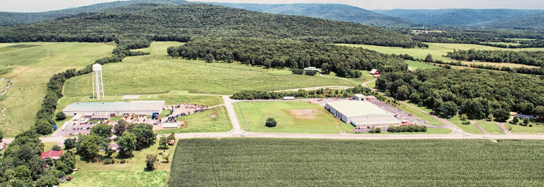 Aerial view of rural landscape with two large industrial buildings, a water tower, open fields, trees, and forested rolling hills in the background under a clear sky.
