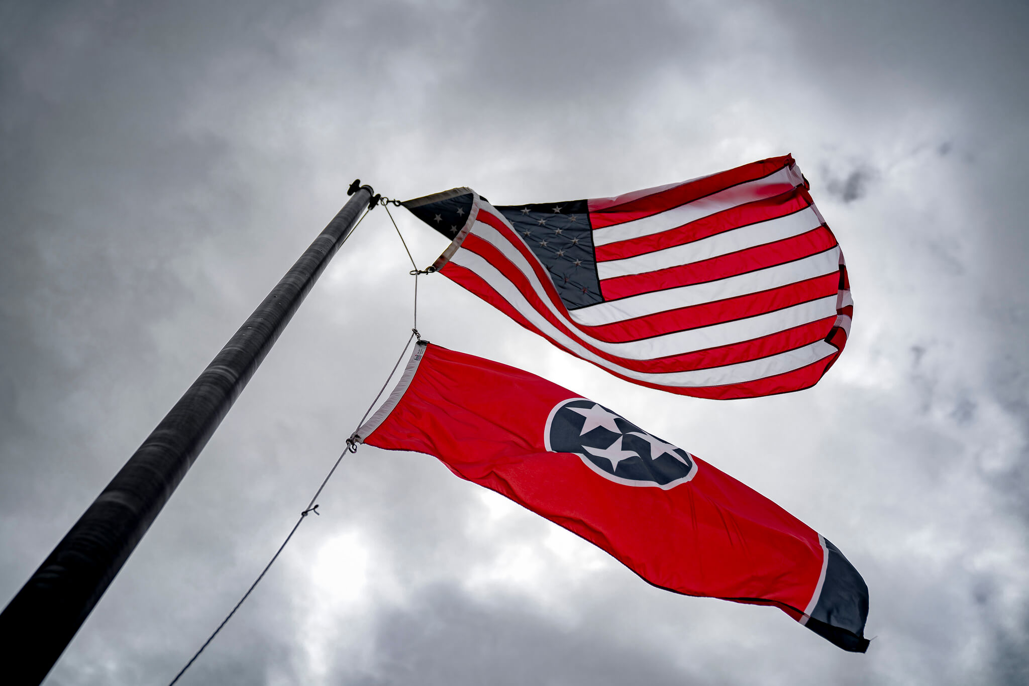 Two flags wave in the wind on a flagpole against a cloudy sky: the United States flag above the Tennessee state flag, which is red with a blue circle and three white stars.