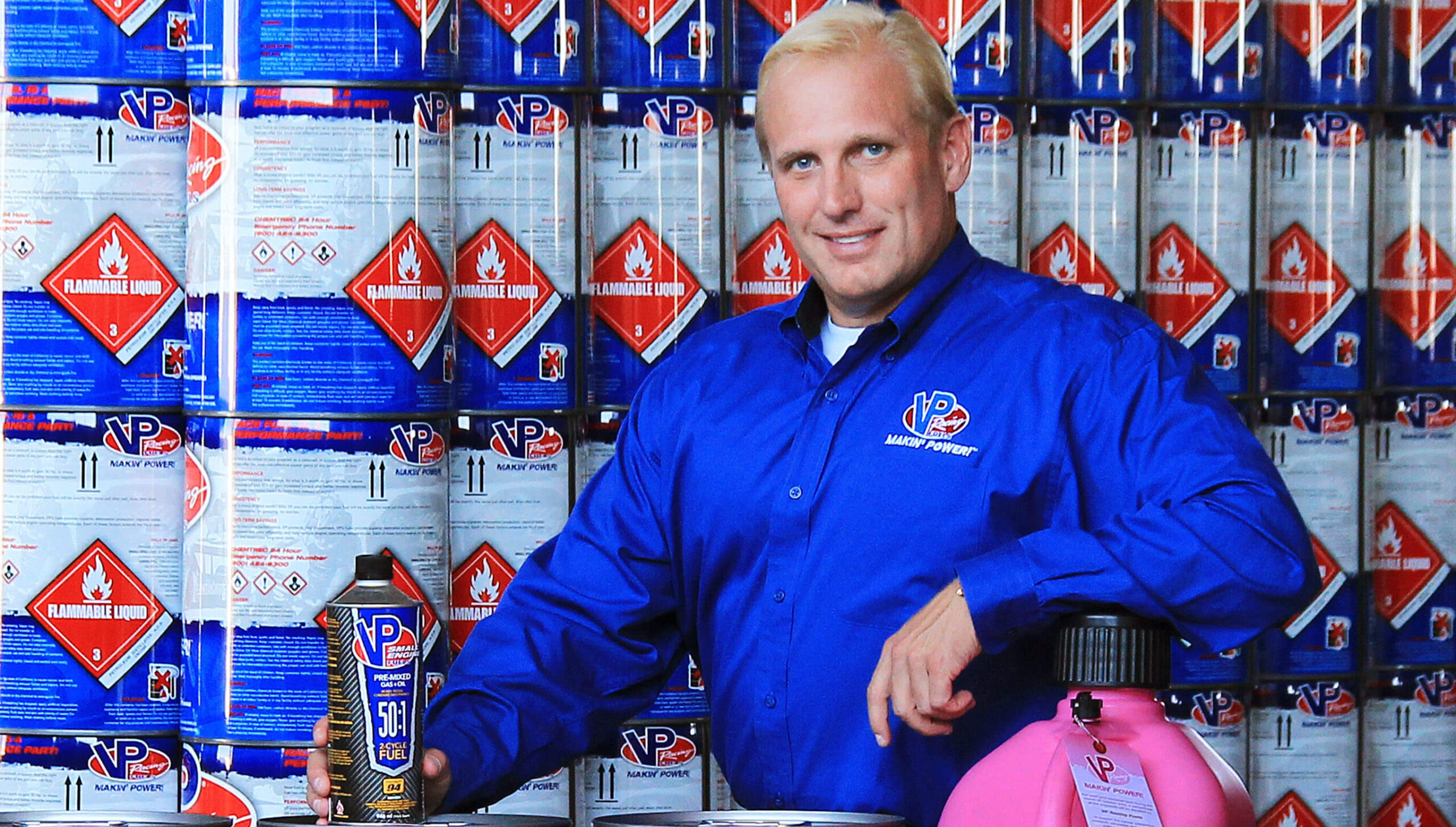 A person in a blue VP-branded shirt stands in front of stacked fuel cans with warning labels, holding a VP Racing Fuels product and resting a hand on a large pink container.