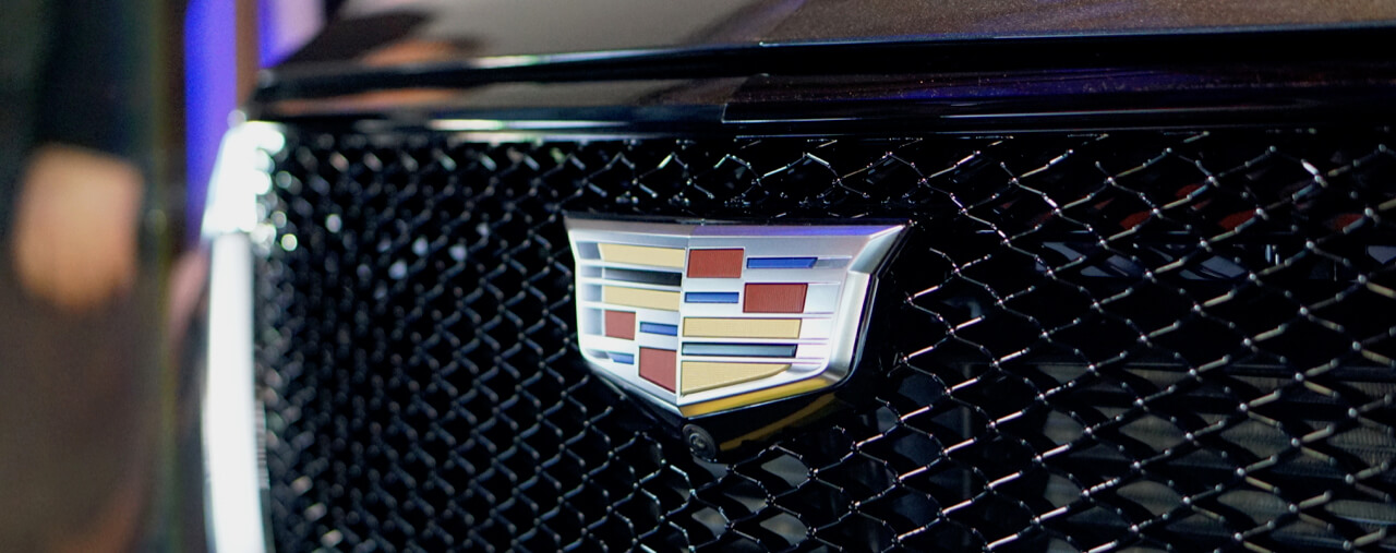 Close-up of a Cadillac logo on the front grille of a black vehicle, featuring a shiny, detailed emblem with red, yellow, blue, and silver colors on a black mesh background.