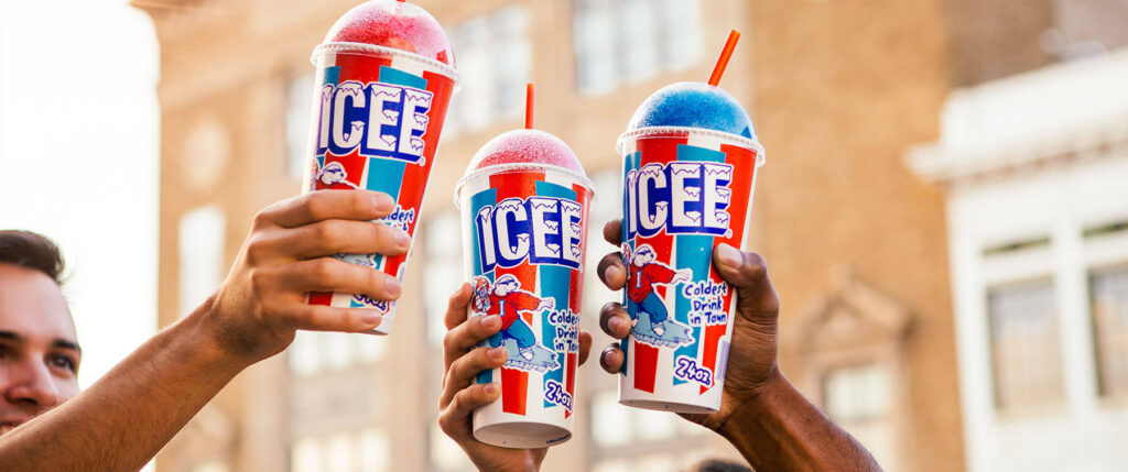 Three people holding up large ICEE cups with red and blue slush drinks, each with a straw, against a blurred outdoor background with buildings.