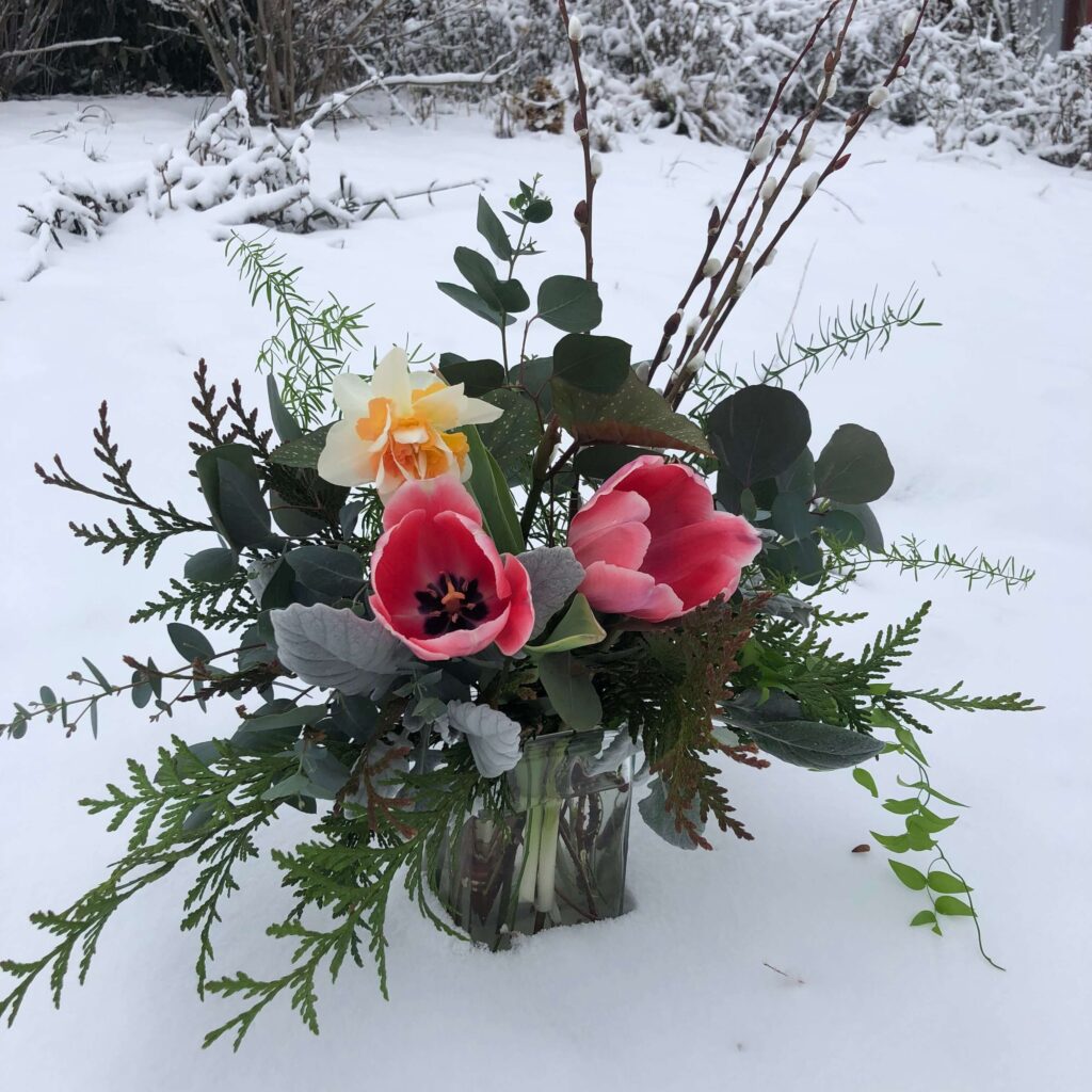 A glass vase with pink tulips, a yellow-white daffodil, and assorted green foliage sits on fresh snow in a snowy outdoor setting with bare branches in the background.
