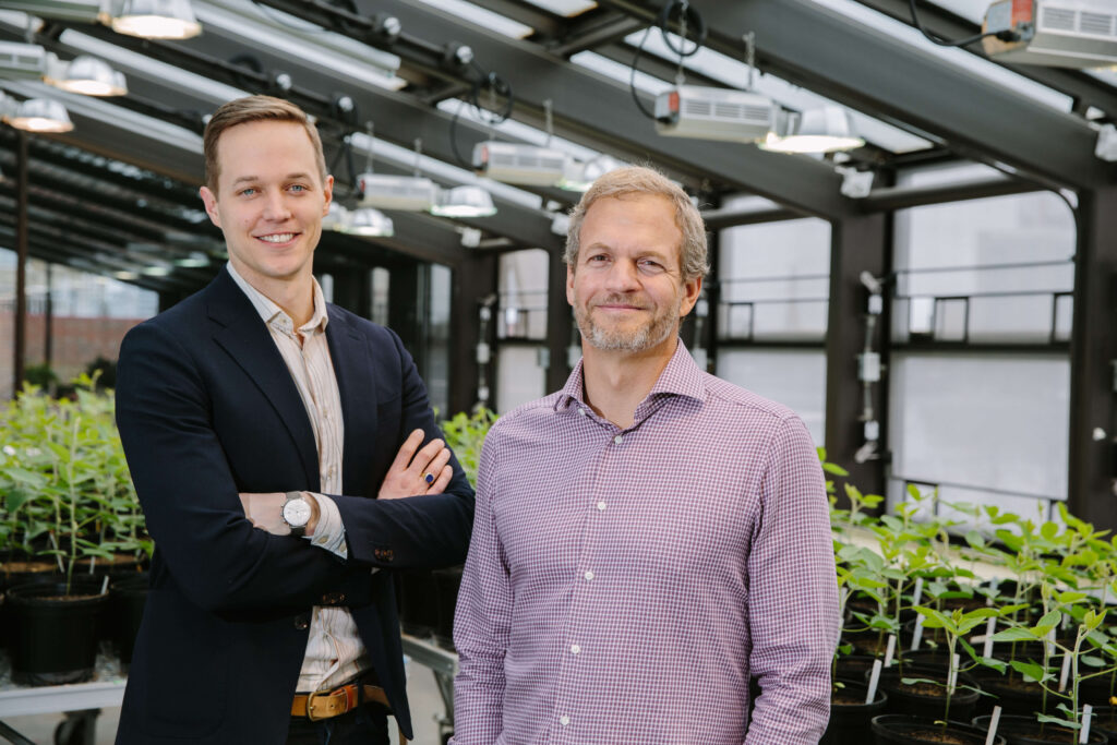 Two men stand smiling in a greenhouse surrounded by potted plants. One wears a dark blazer with arms crossed, while the other wears a light checkered shirt. Sunlight streams in through the glass roof above.