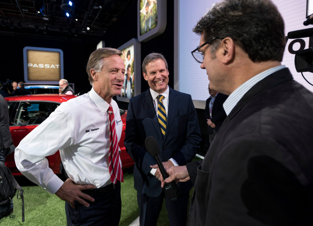 Three men in business attire talk at an event next to a red car. One man holds a microphone, suggesting an interview. Signs in the background display "PASSAT" and images of cars, indicating an automotive event.