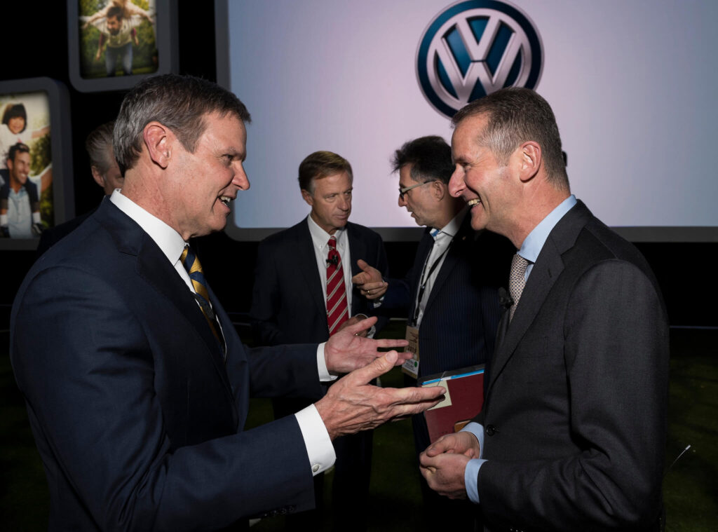 Two men in suits are engaged in conversation at an event, smiling at each other. Three other men in suits are talking in the background. A large Volkswagen logo is displayed behind them on a screen.