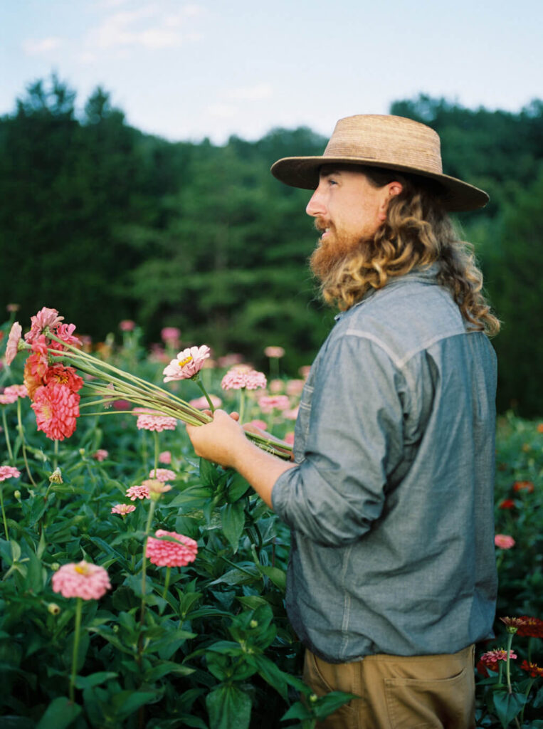 A person with long curly hair, a beard, and a straw hat stands in a flower field, holding pink flowers and looking to the side. Trees and greenery are visible in the background under a blue sky.