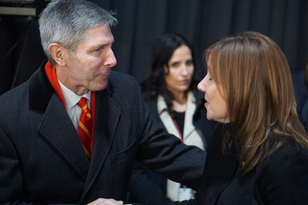 A man in a suit with a red and yellow striped scarf shakes hands and speaks with a woman in a black coat, while another woman stands in the background, looking on.