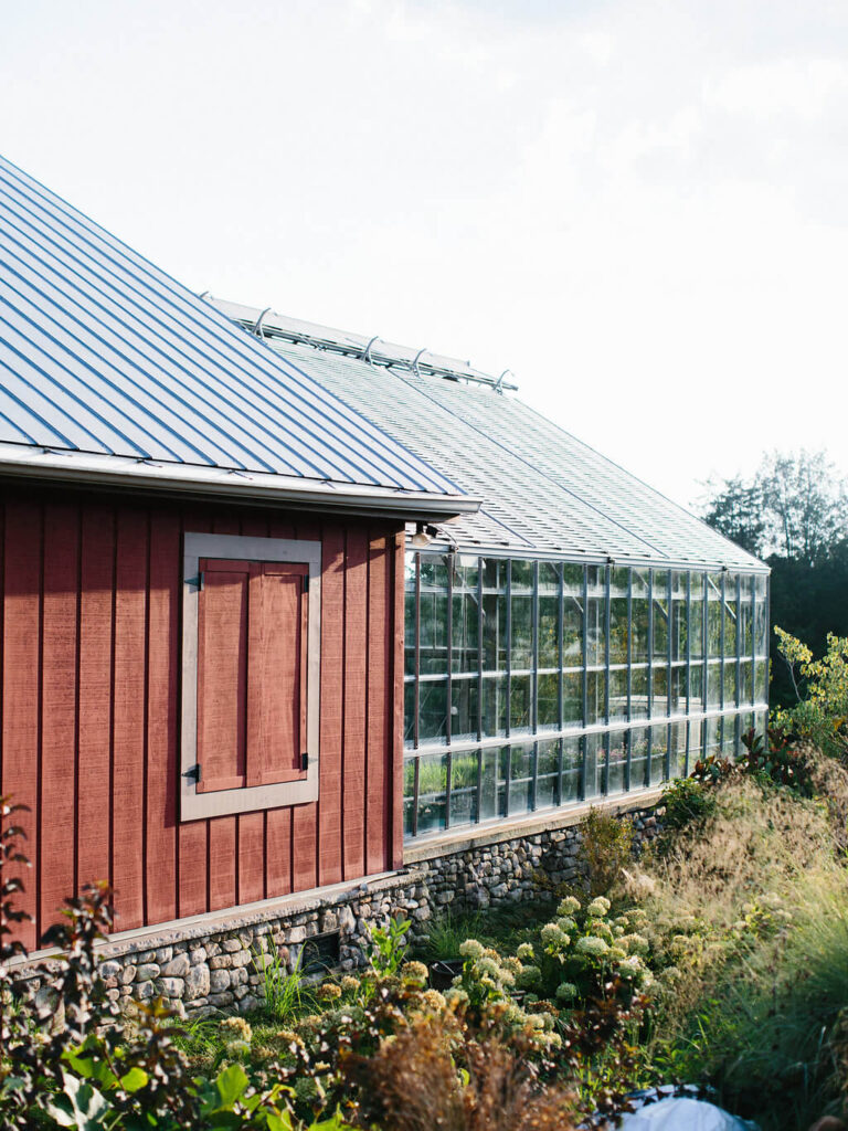 A red wooden building with a closed window is attached to a glass greenhouse. Green plants and shrubs grow alongside the stone foundation under a bright, clear sky.