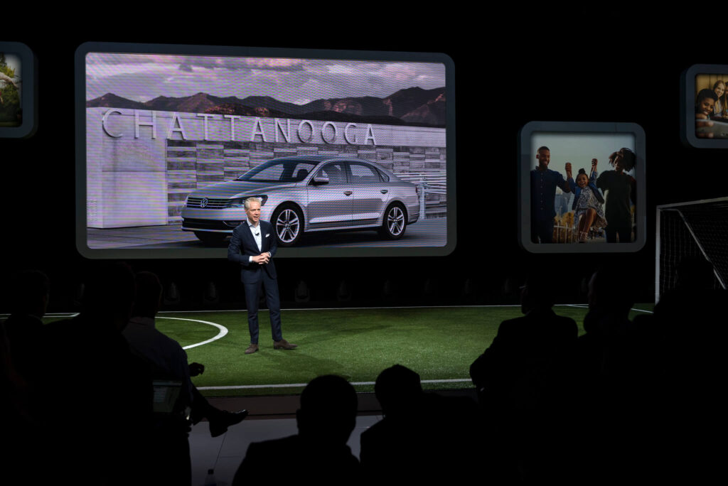 A man in a suit speaks on stage in front of a large screen displaying a car and the word "Chattanooga," while an audience watches in a dimly lit room.