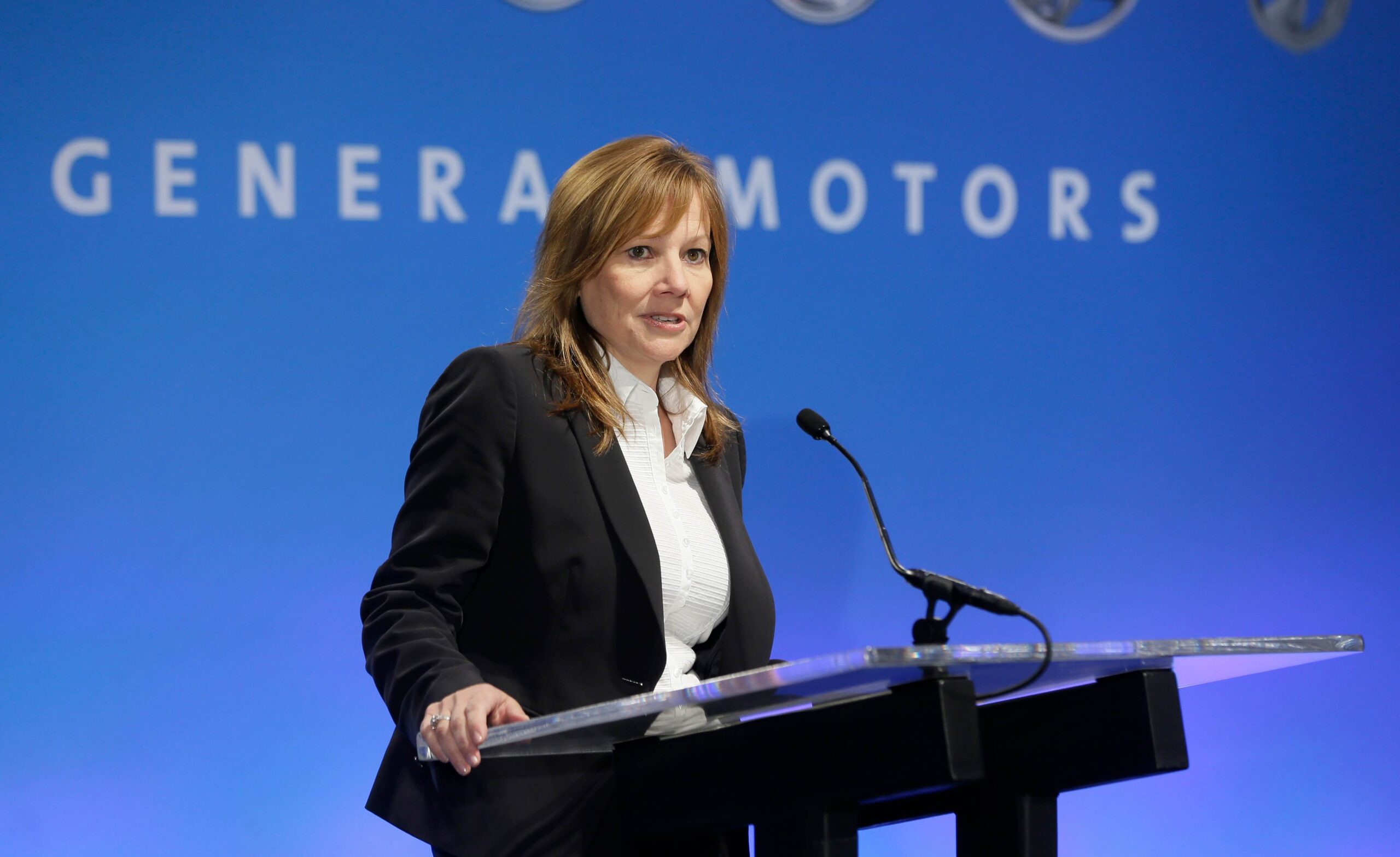 A woman in a business suit speaks at a podium with a microphone in front of a blue background displaying the words "GENERAL MOTORS.