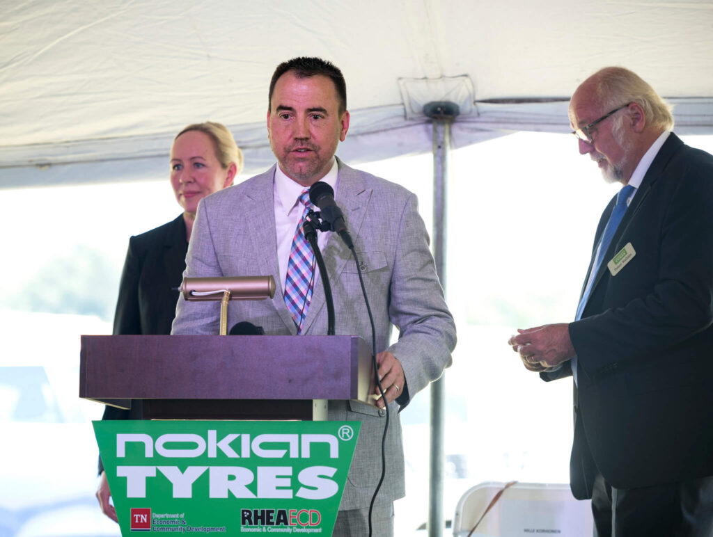 A man in a light gray suit speaks at a podium with a "Nokian Tyres" sign, while a woman and another man stand nearby under a white tent. All appear to be at an outdoor event or ceremony.