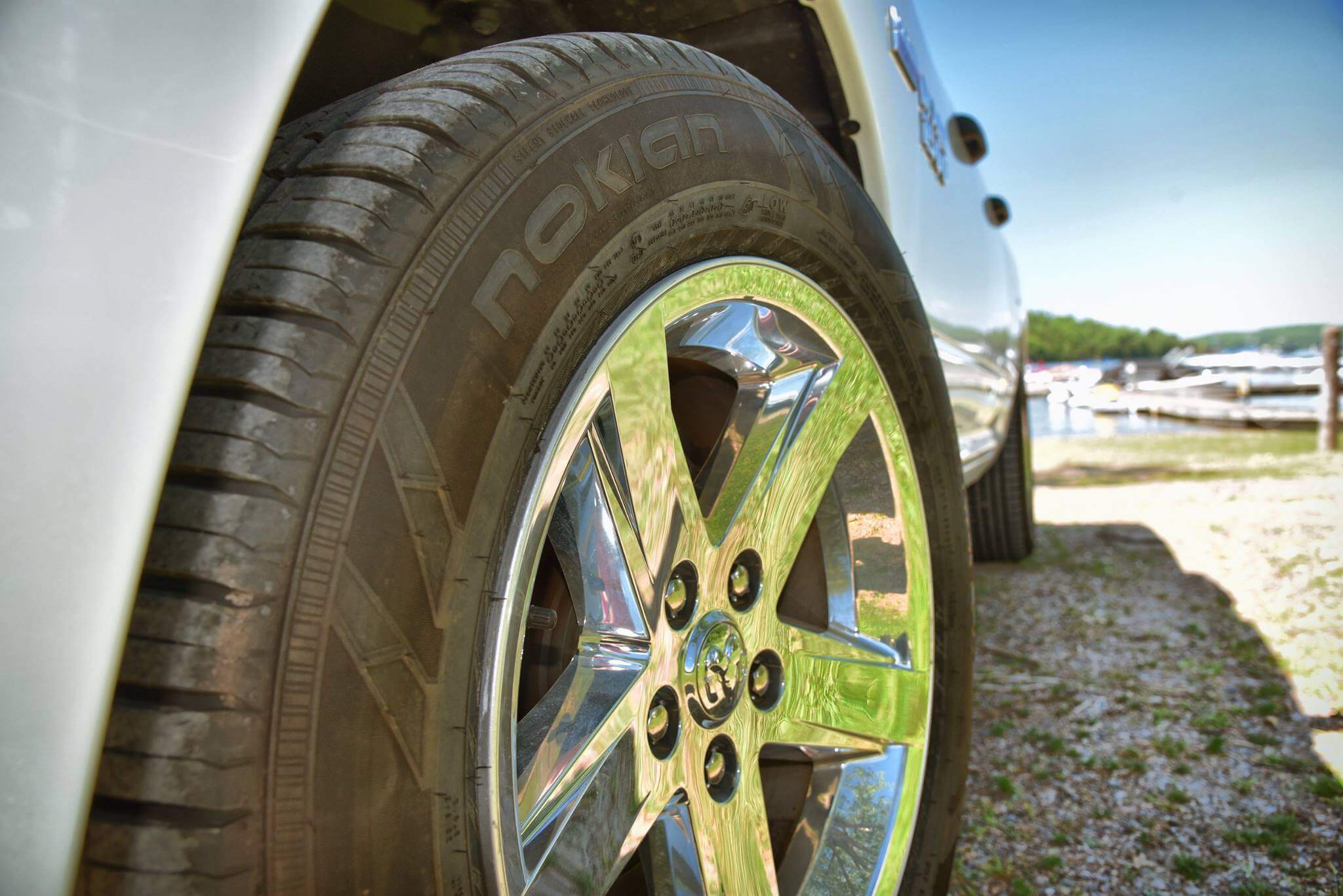 Close-up view of a shiny chrome wheel and Nokian tire on a silver vehicle, parked on a gravel surface near a body of water with boats and a tree-lined shore in the background.