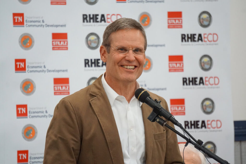 A man in a brown blazer and white shirt smiles while speaking at a microphone, standing in front of a backdrop with logos for the Tennessee Department of Economic & Community Development and RHEA ECD.