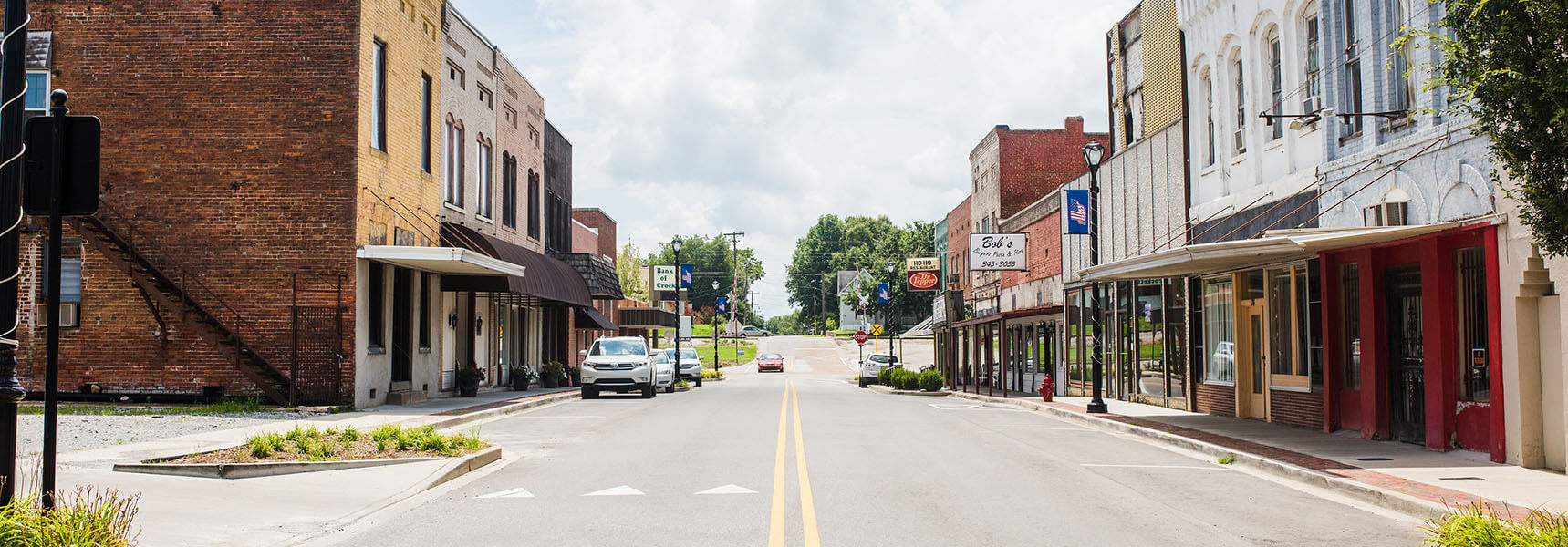 A quiet small-town main street with brick and painted buildings, shopfronts, parked cars, and a double yellow line running down the center under a partly cloudy sky.