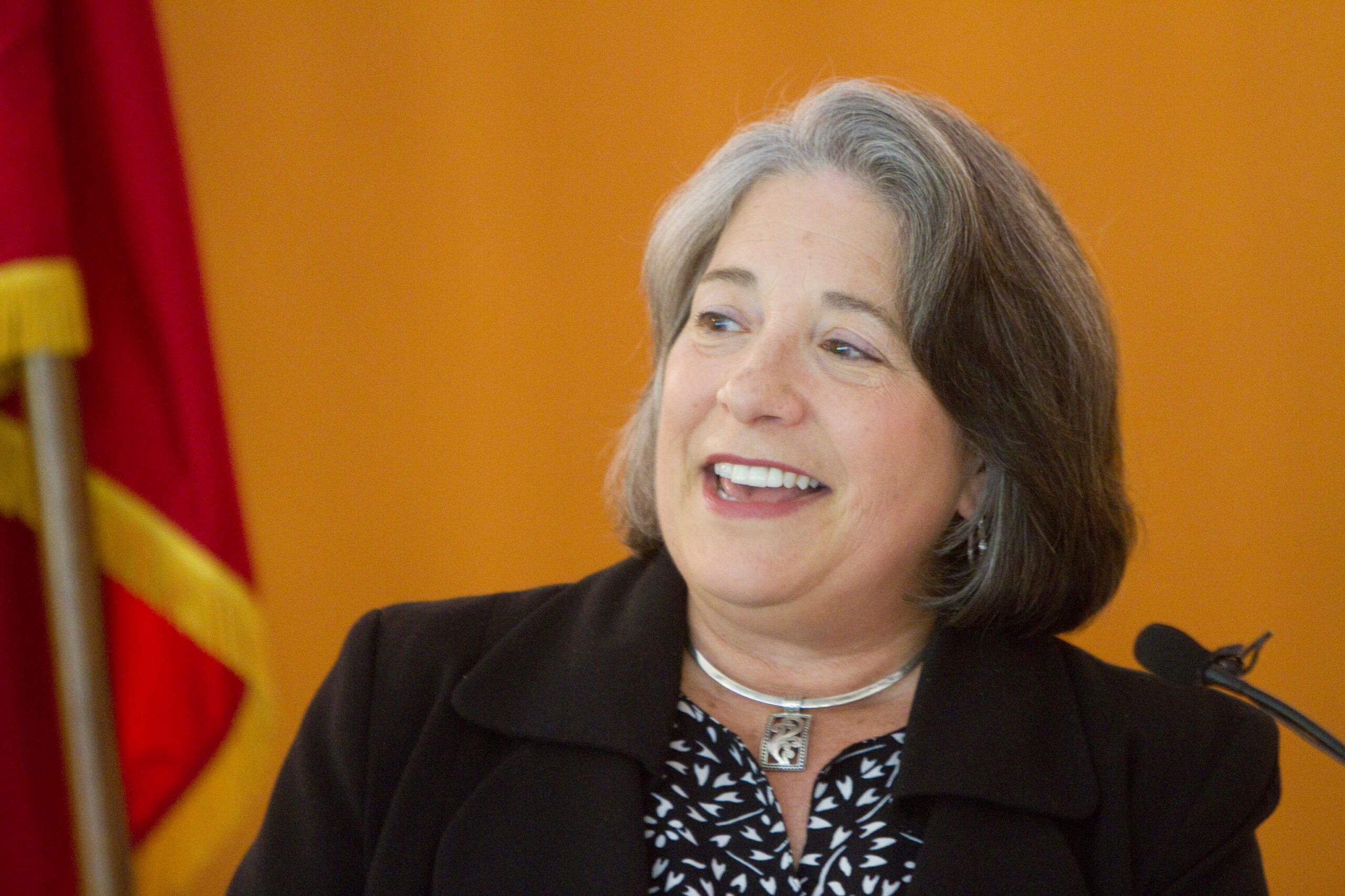 A woman with shoulder-length gray hair is smiling while speaking. She is wearing a black blazer, patterned top, and a necklace. An orange background and part of a red and yellow flag are visible behind her.
