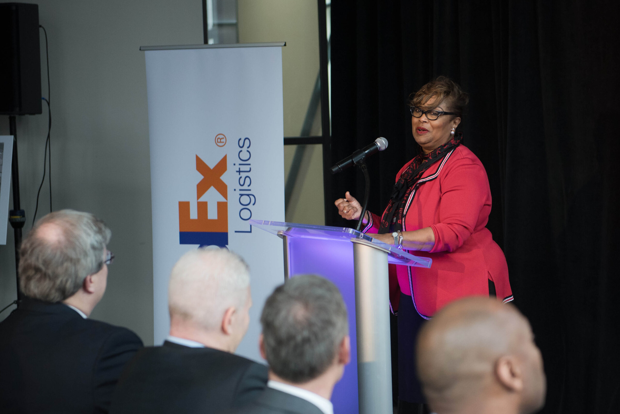 A woman in a red blazer speaks at a podium with a microphone in front of an audience. A banner with the logo "Ex Logistics" is visible in the background.