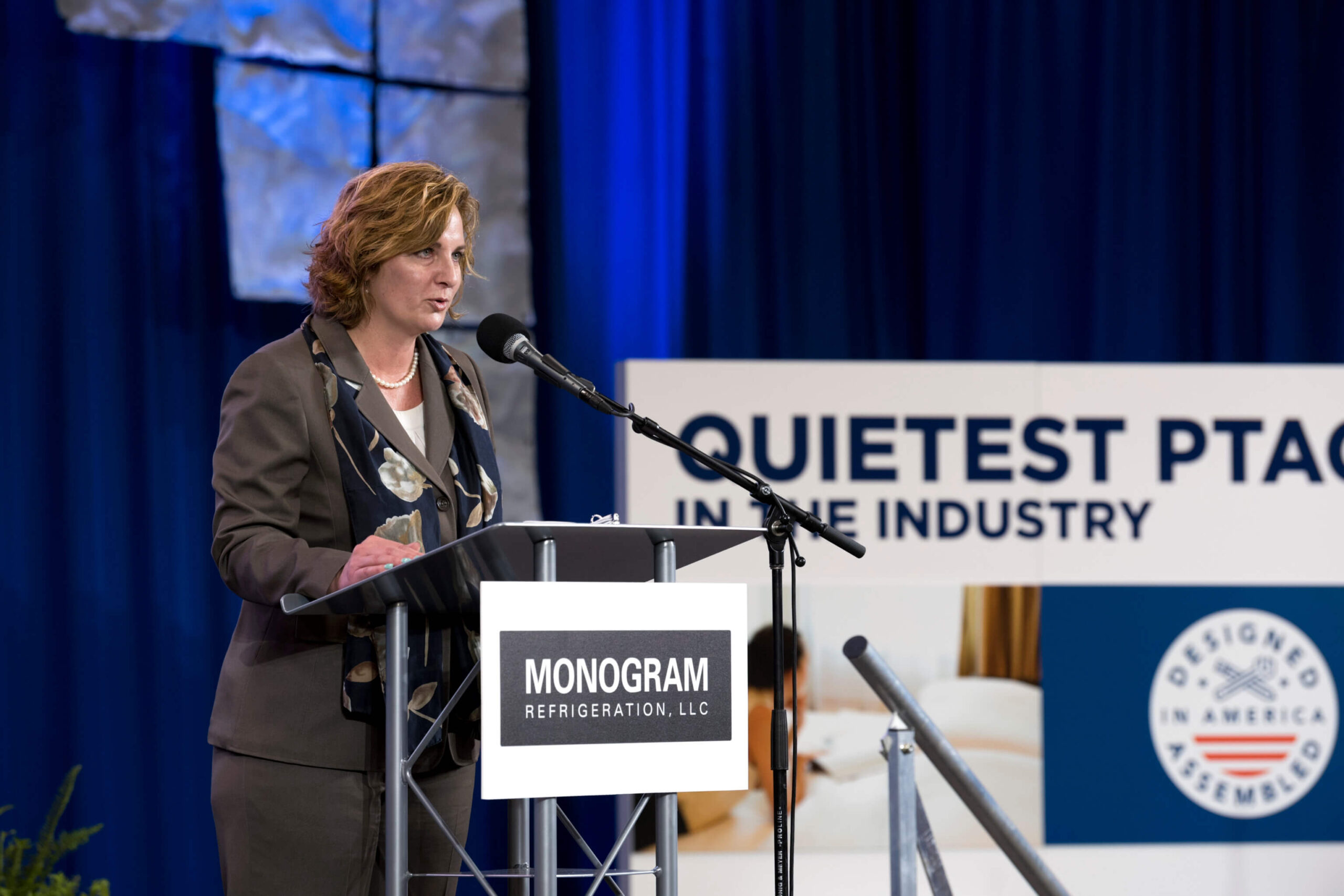 A woman in a suit speaks at a podium with a microphone. Behind her is a blue sign that reads, "QUIETEST PTAC IN THE INDUSTRY" and a logo for Monogram Refrigeration, LLC. The background is blue with metal accents.