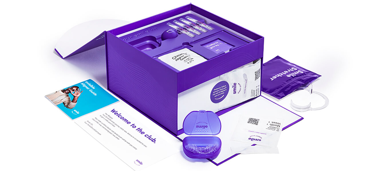 A purple SmileDirectClub orthodontic starter kit is displayed with dental aligners in a case, impression tools, an instruction booklet, a welcome card, and other accessories arranged neatly around the box.