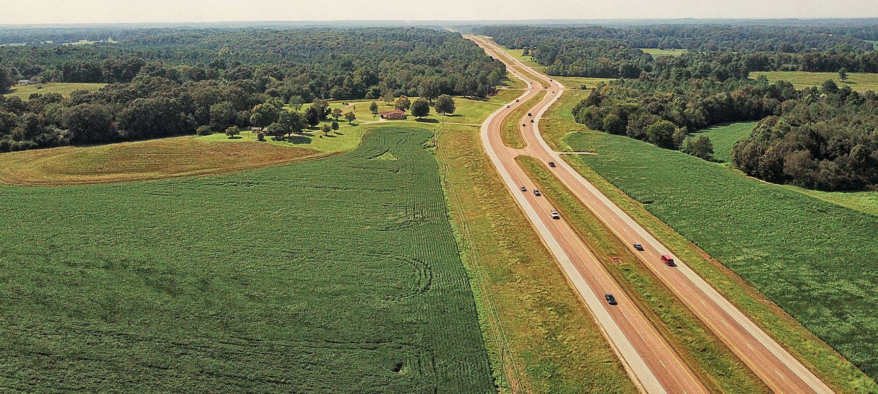 Aerial view of a rural highway with cars traveling in both directions, surrounded by green fields and scattered trees under a clear sky.