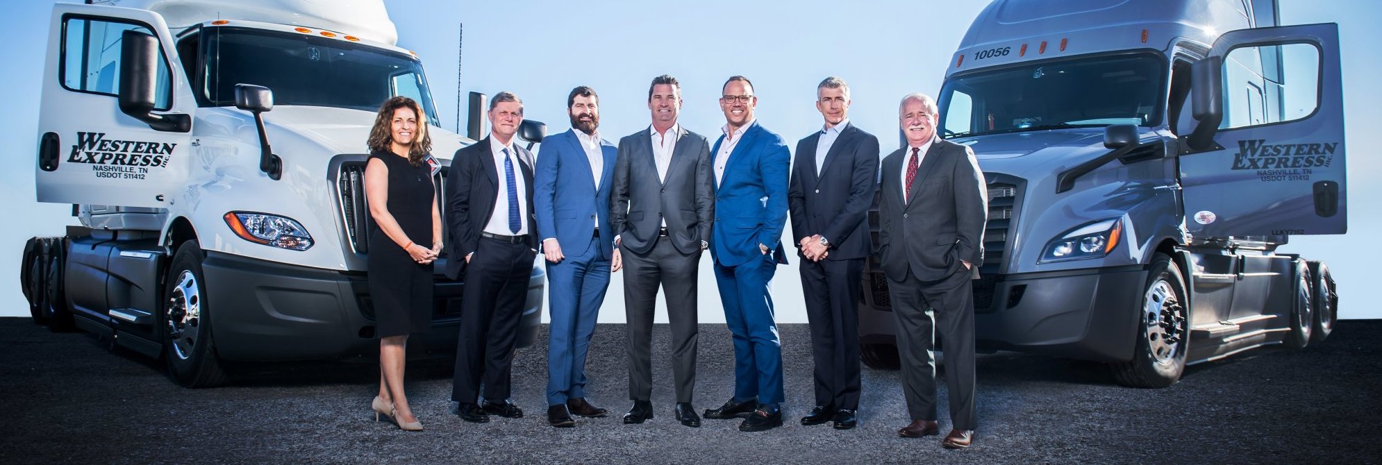 Seven professionally dressed people stand smiling between two Western Express semi-trucks parked facing each other on a paved surface under a clear blue sky.