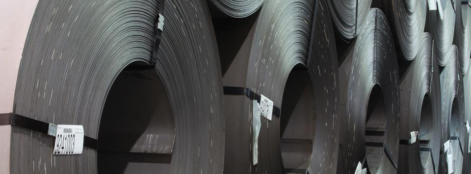 Large rolls of sheet metal are stacked vertically in a row inside an industrial facility, each with labels attached to the outer edges.