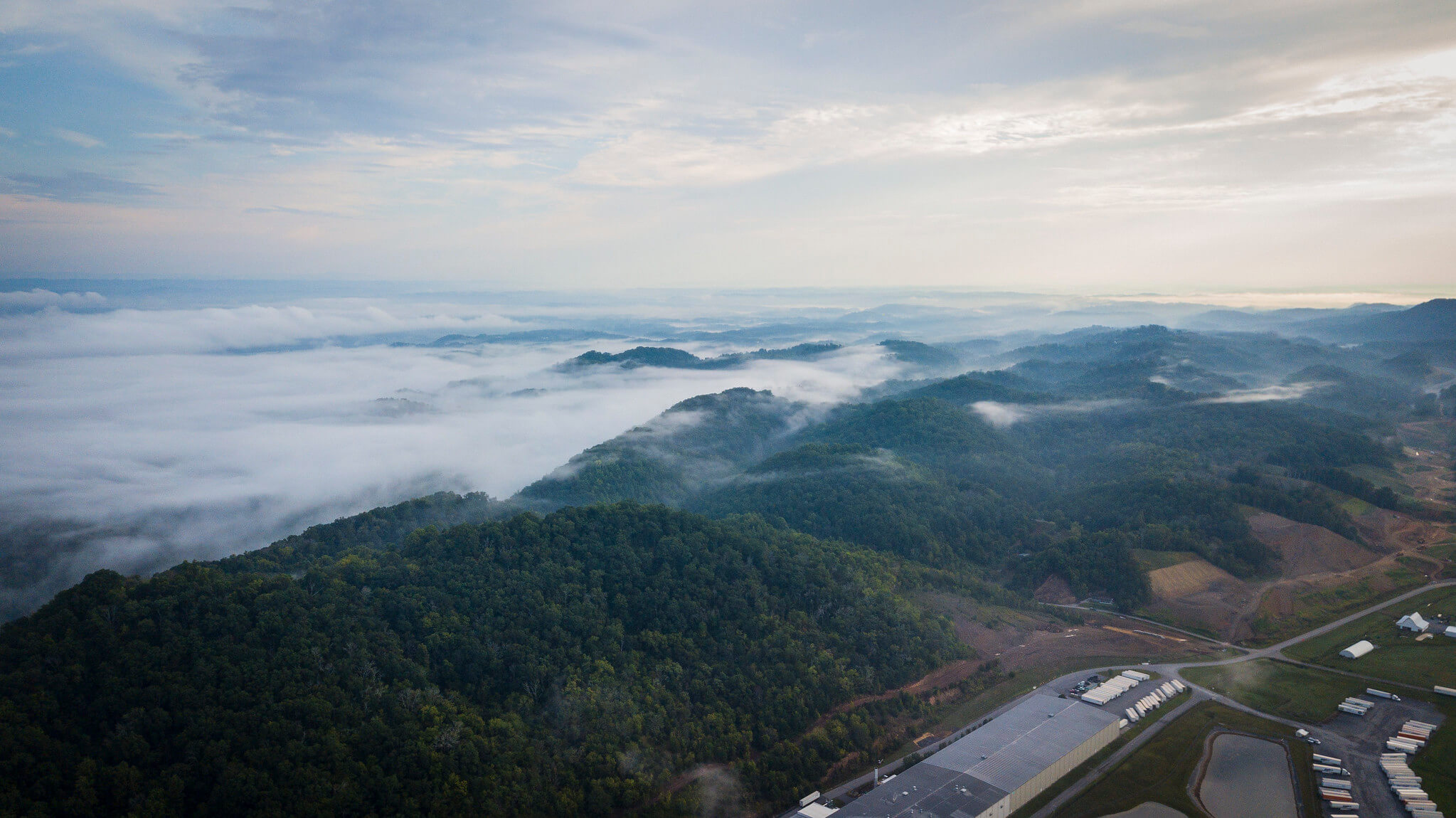 Aerial view of rolling green hills covered with trees and low-lying morning fog, under a cloudy sky. Industrial buildings and roads are visible in the lower right corner.