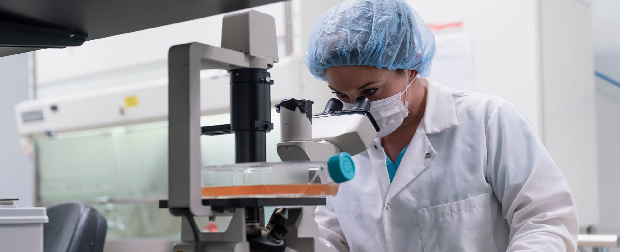 A scientist wearing a lab coat, hair cover, and face mask looks into a microscope in a modern laboratory setting. Lab equipment and safety gear are visible in the background.