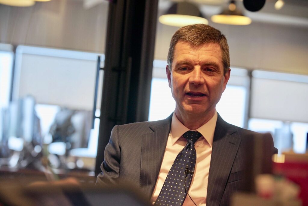 A man in a suit and tie sits indoors, speaking or listening, with blurred office windows and lights in the background.