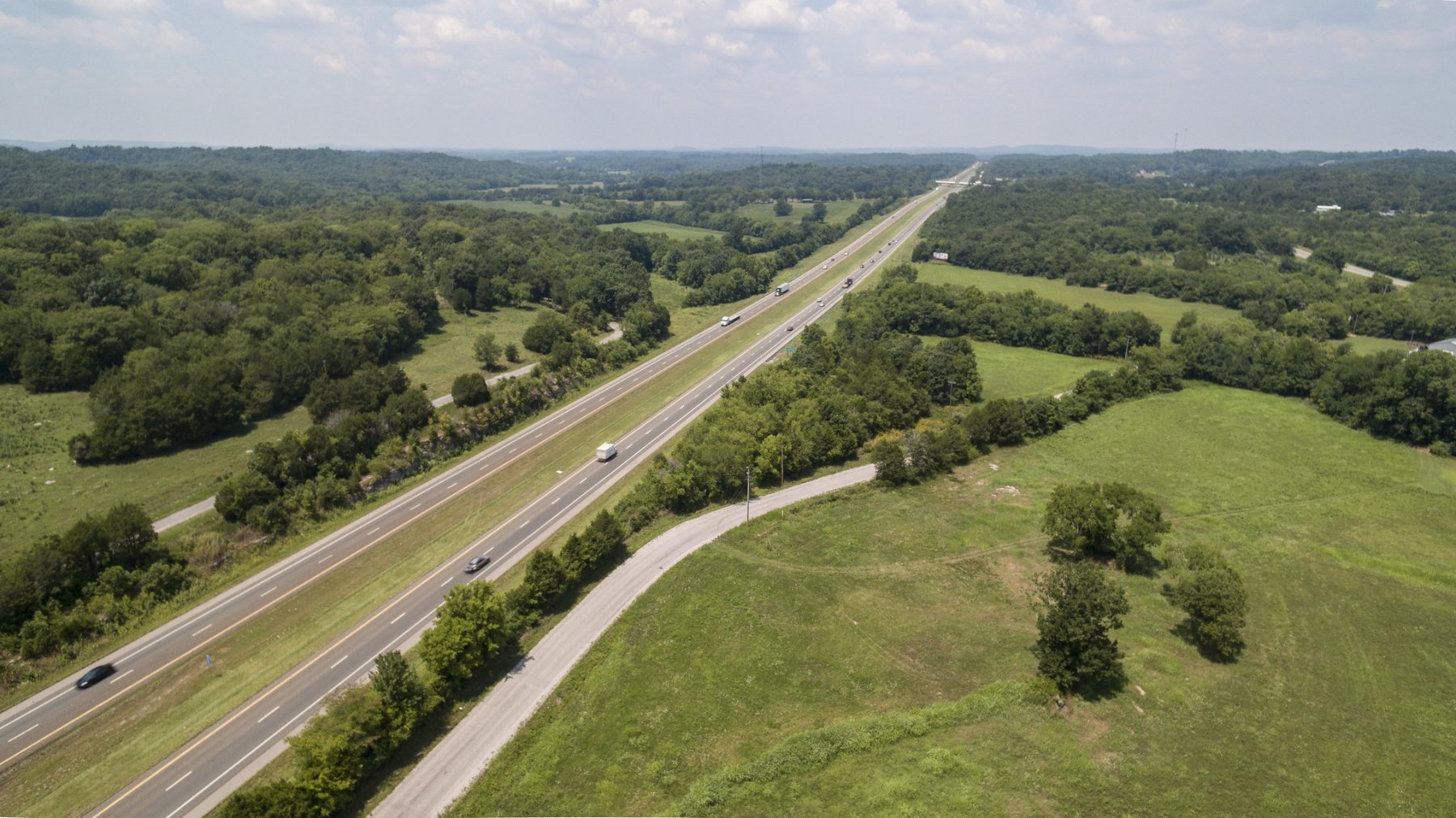 Aerial view of a highway running through green fields and forests, with a few cars traveling in both directions under a lightly cloudy sky.