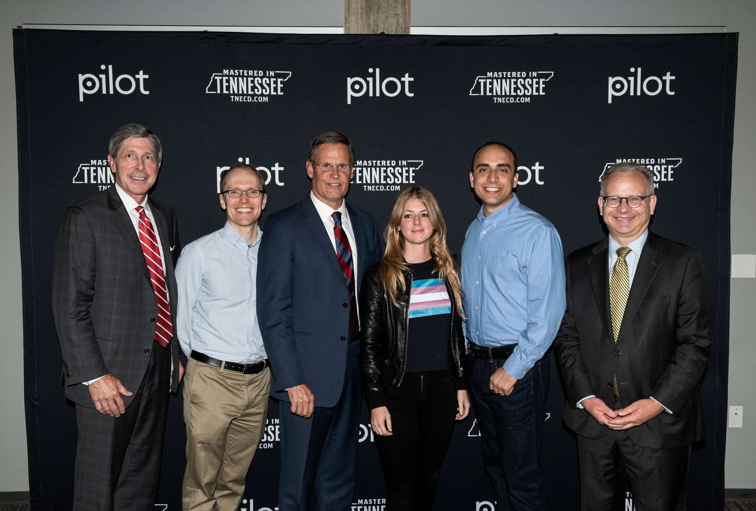 Six people stand in front of a black backdrop with "Pilot" and "Mastered in Tennessee" logos, posing and smiling for a group photo. Most are in business attire; one person wears a black jacket and colorful T-shirt.