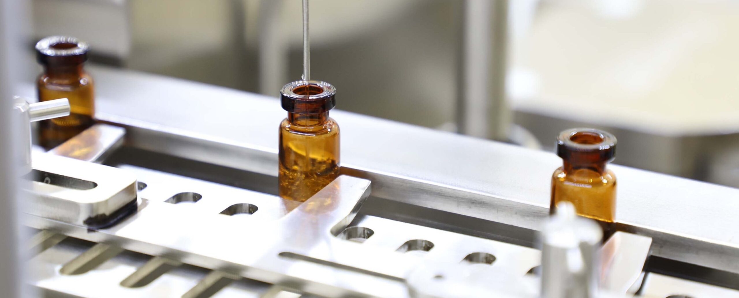 Close-up of a pharmaceutical production line where a needle fills small brown glass vials with liquid. The vials are aligned in a metal tray, part of an automated filling process.