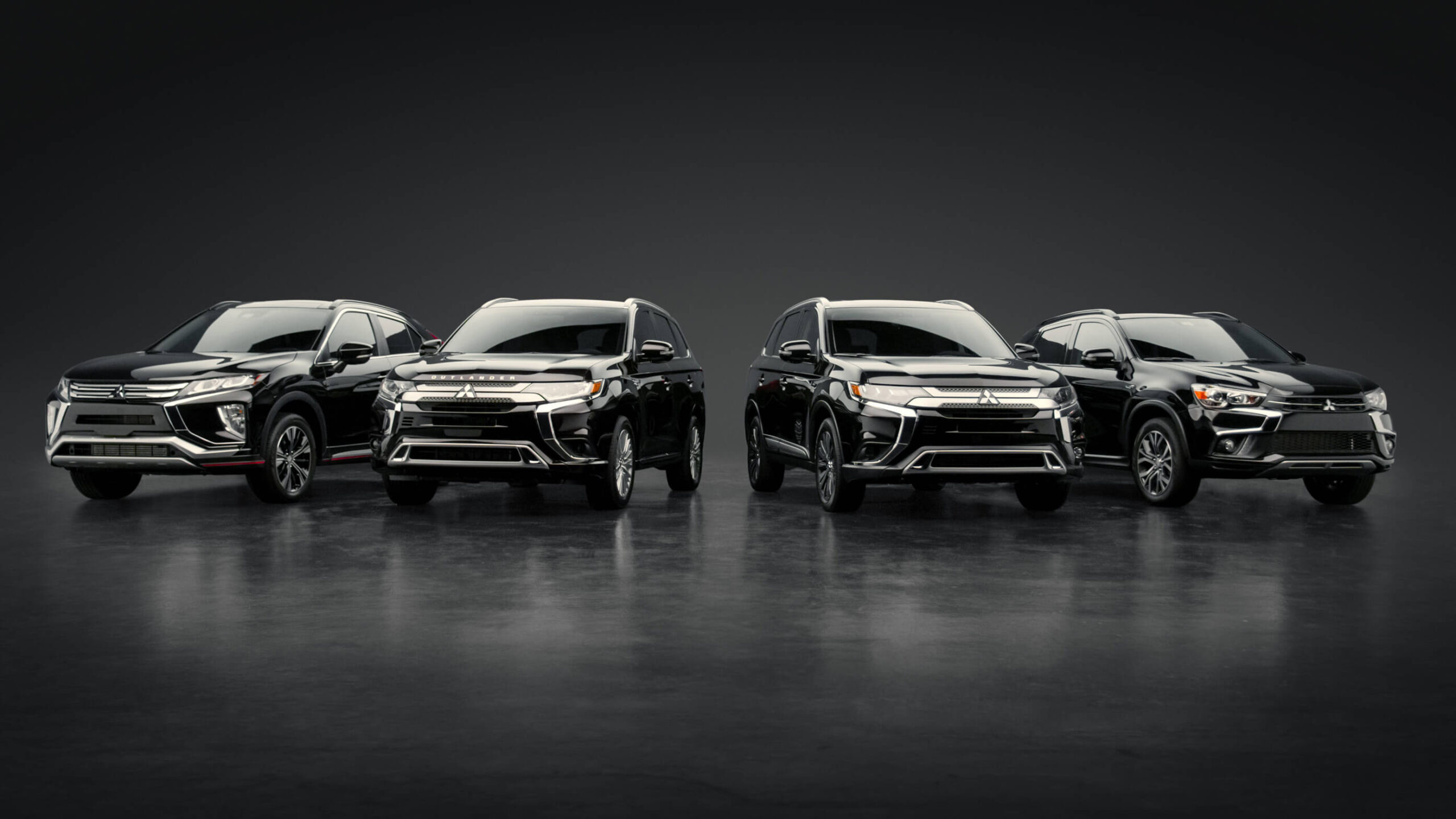 Four black Mitsubishi SUVs are lined up side by side on a glossy, dark surface with a dark, minimal background, facing the camera. The vehicles are well-lit and display slight design differences.