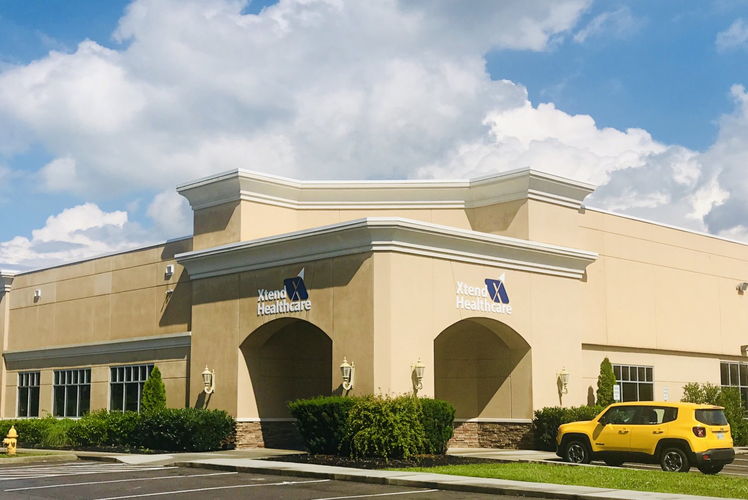 A tan, one-story building with "Xlendi Healthcare" signs on two walls, surrounded by bushes and a parking lot. A bright yellow SUV is parked near the front entrance under a partly cloudy sky.
