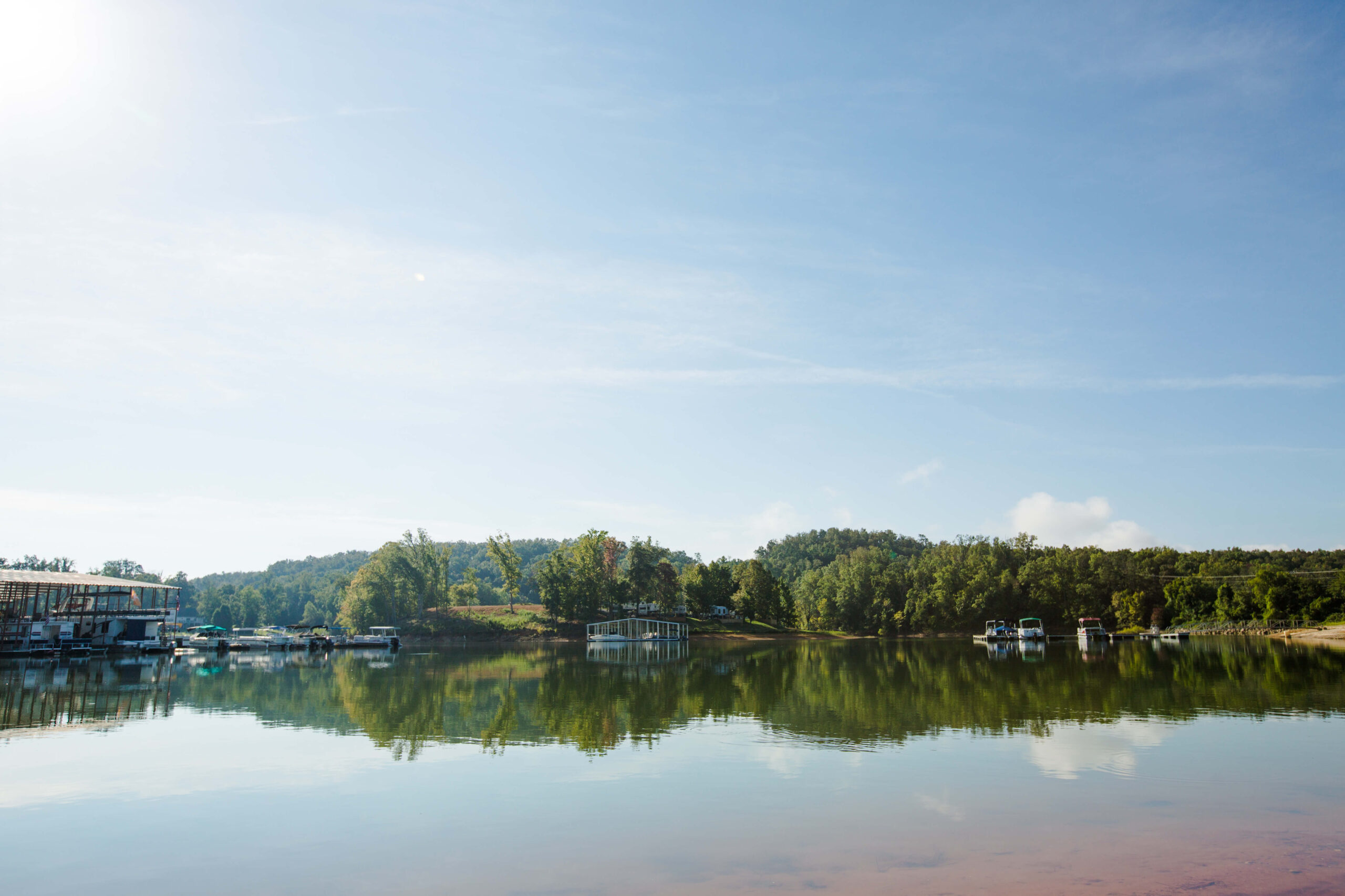 A calm lake with clear reflections of trees and boats along the shore under a bright blue sky, with sunlight streaming from the top left corner.