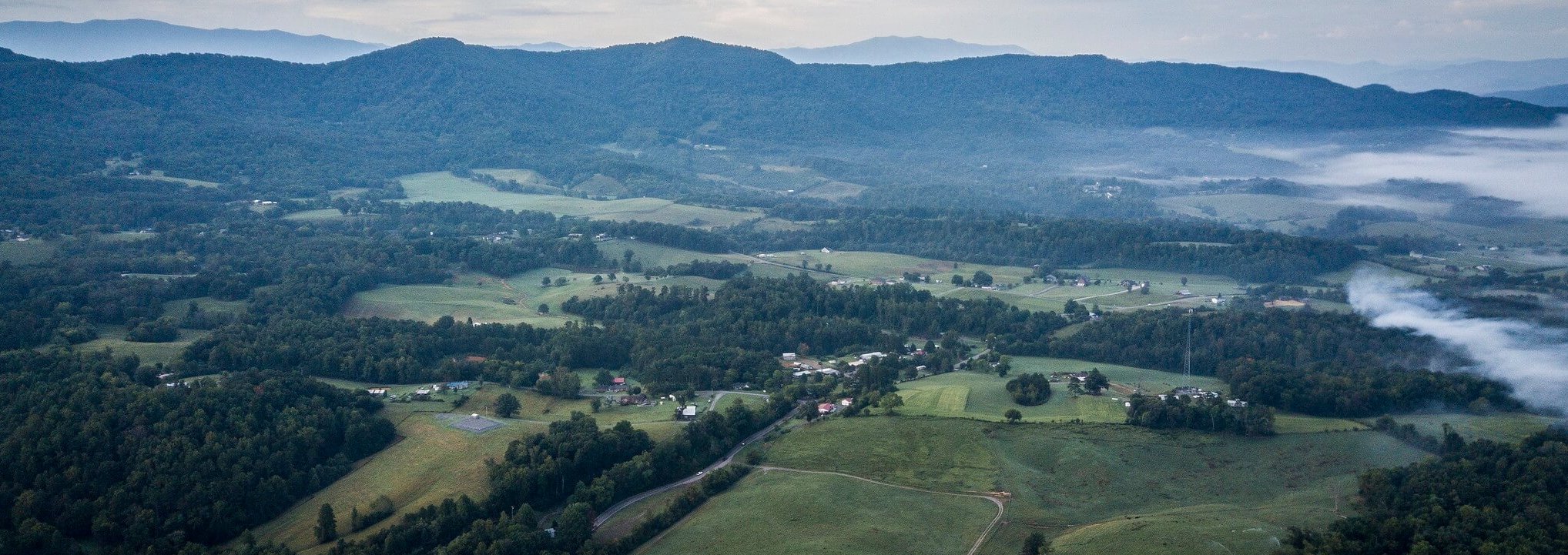 Aerial view of a rural landscape with scattered houses, green fields, winding roads, dense forests, and rolling hills, with mist and distant mountains under a cloudy sky.