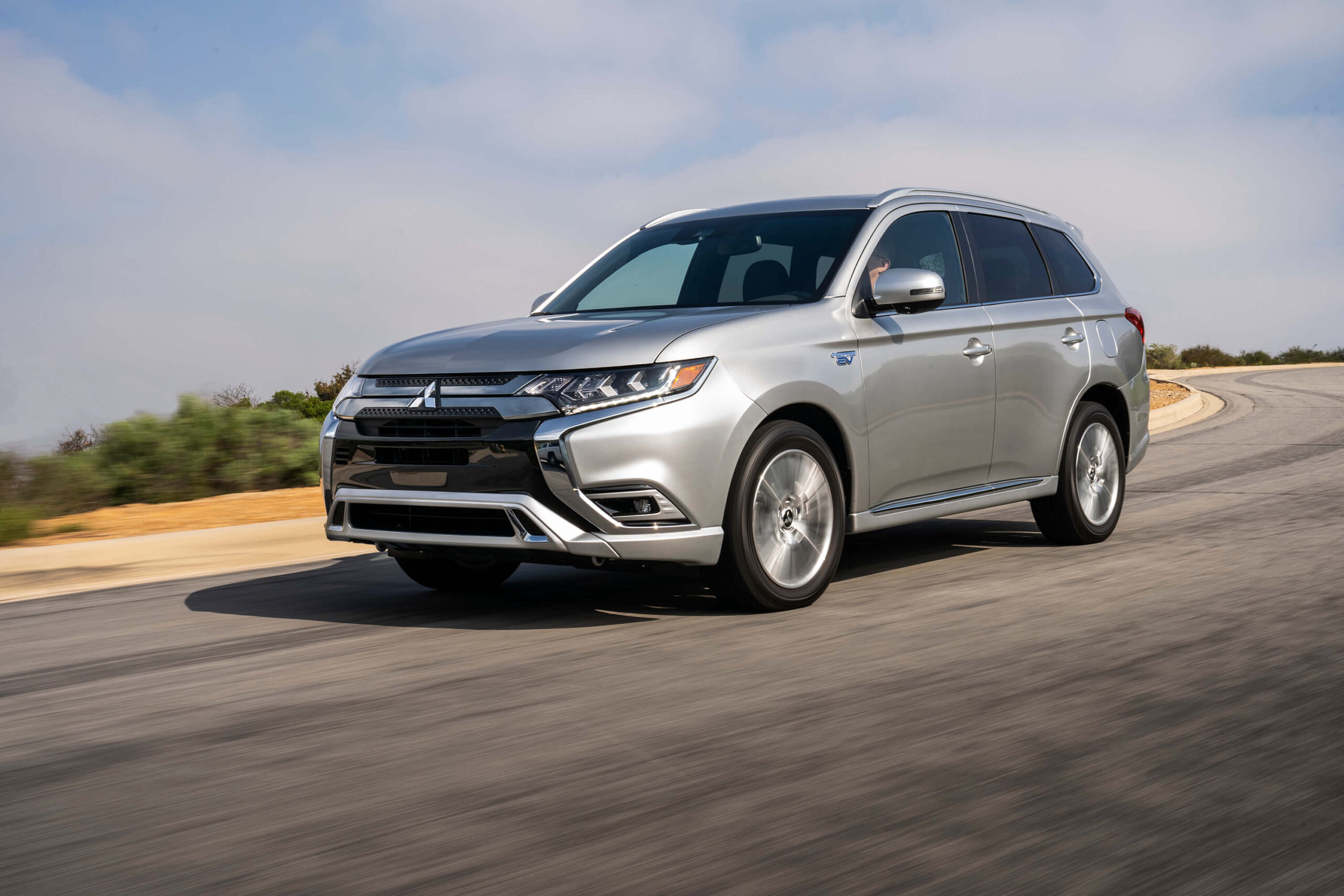 A silver Mitsubishi Outlander SUV drives on a curved road with a partly cloudy sky and greenery in the background.