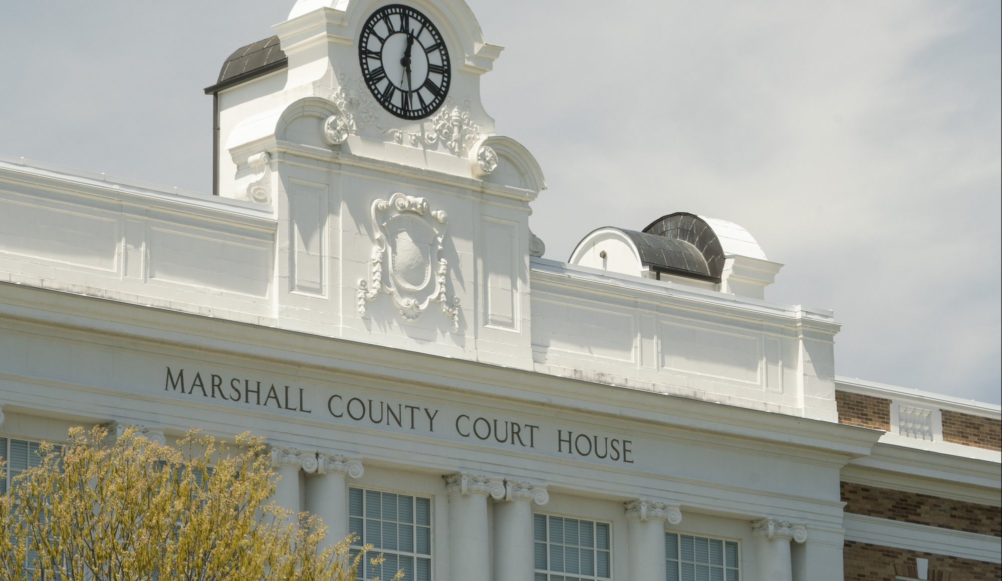 White facade of the Marshall County Court House with a black and white clock and ornate architectural details above tall windows, set against a partly cloudy sky.