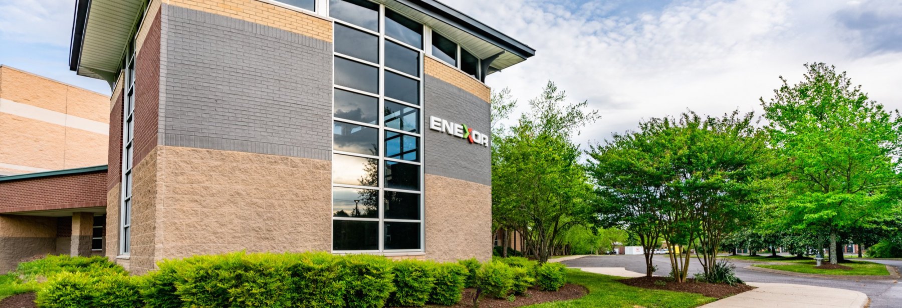 A modern office building with large windows and the logo "ENEROR" on the exterior, surrounded by green bushes and trees under a partly cloudy sky.
