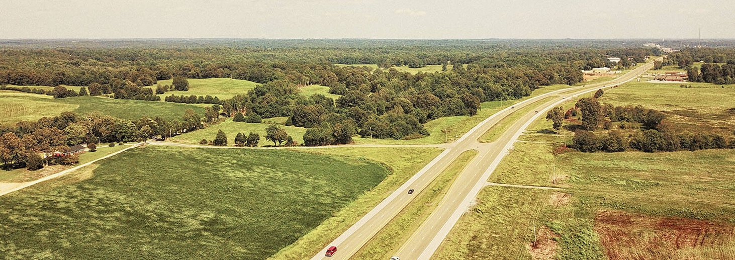 Aerial view of a highway cutting through green fields and patches of trees on a sunny day, with a few cars traveling along the road and rural buildings visible in the distance.