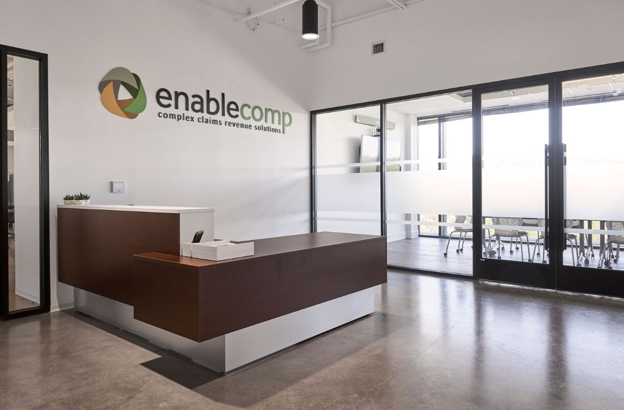 Modern office reception area with a sleek wooden desk, a small plant, and the "enablecomp" logo with the tagline "complex claims revenue solutions" on the wall. Large glass doors lead to a conference room.