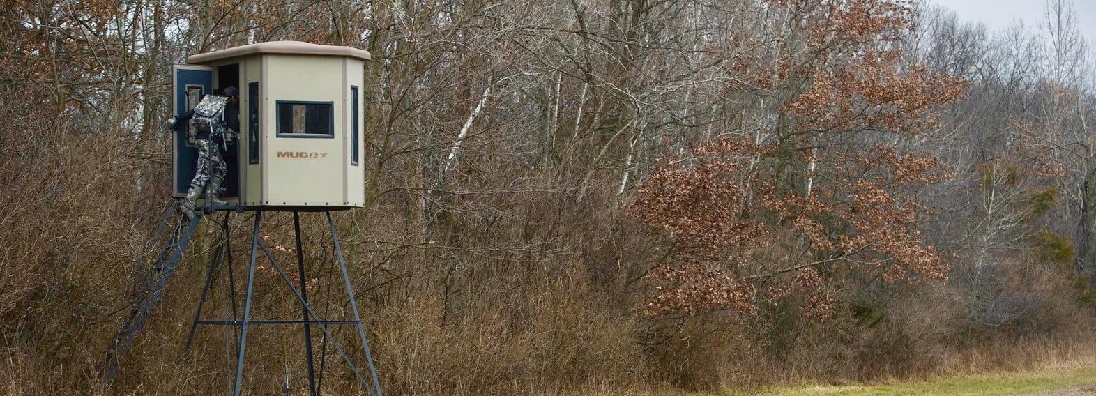A person in camouflage climbs into a raised hunting blind on metal stilts at the edge of a field, with leafless trees and dry grass in the background.