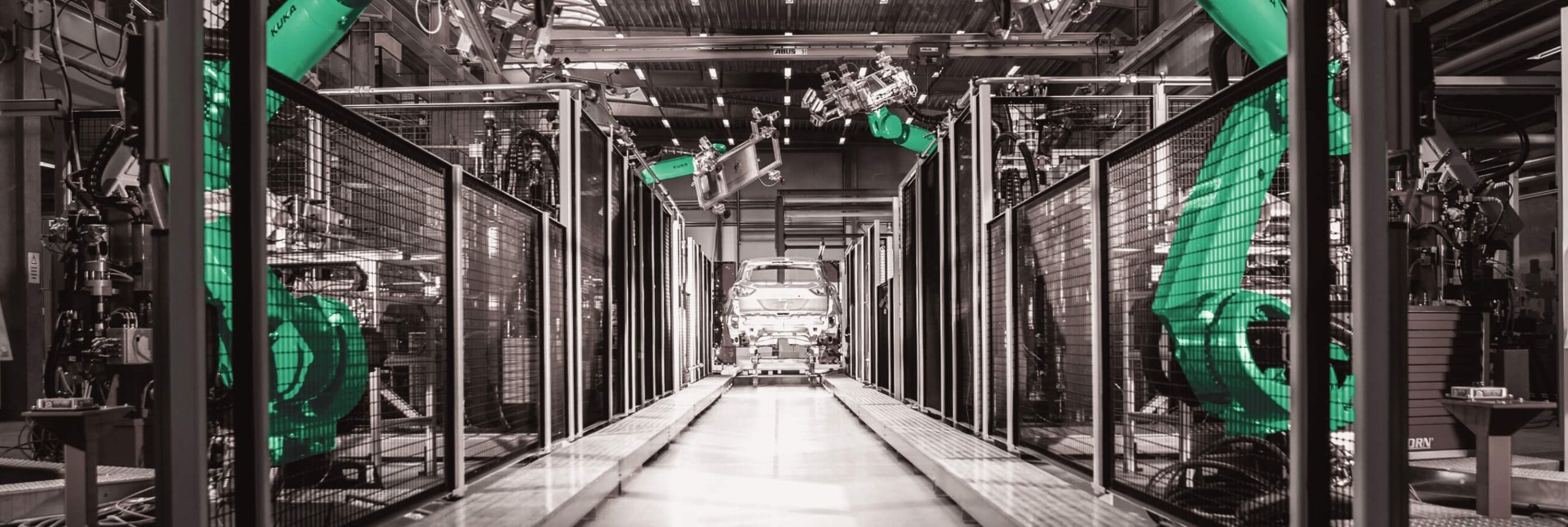 Robotic arms assemble a car on an automated production line inside a modern factory. The image has a monochrome tone with green highlighted robots and features safety barriers along the sides.